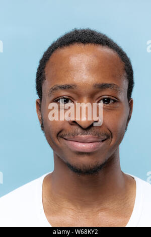 Close up portrait of young african-american man in white shirt on blue background. Human emotions, facial expression, ad, sales concept. Looks calm. Looking at camera. Stock Photo