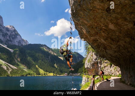 GOSAU, AUSTRIA - JULY 4 2019: Man climbing on Laserer alpin via ferrata over Vorderer Gosausee lake with Grosser Donnerkogel Mountain in background Stock Photo