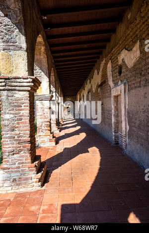 Weathered and worn brick arches form the famous corridors, casting large shadows on red floor tiles at the Mission San Juan Capistrano, California. Stock Photo