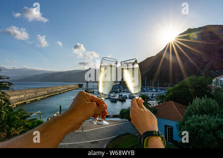 Two hands holding two glasses of champagne in front of a marina during sunset Stock Photo