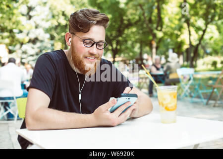 Attractive redhead man with a beard listens to music on a mobile phone while sitting at a cafe table. Smiling male uses a smartphone Stock Photo
