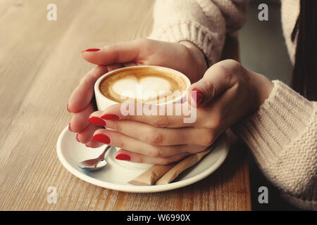 Close-up female hands holding cup with coffee cappuccino with foam with pattern heart. Perfect red gel polish manicure. Wood natural table. Creative c Stock Photo