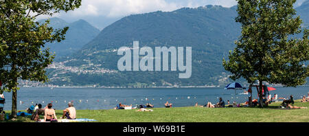 Lake Como, Italy - July 21, 2019: People having rest on beach near lake in summer day. Green trees and Alp mountains on background. Panoramic view.
