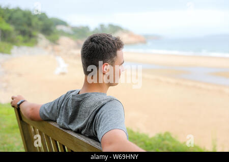 Young man sits on a bench gazing out at the ocean on the coast of Northeast American. Stock Photo