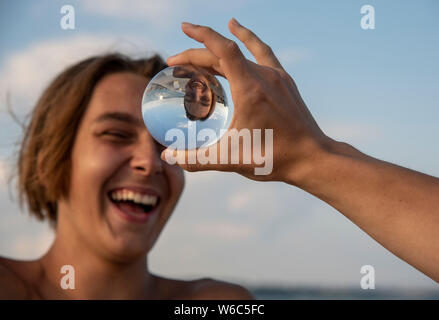 A blurred portrait of a laughing teenager in the background, in the front hand holding a lens ball, which reflects the teenager upside down. Stock Photo