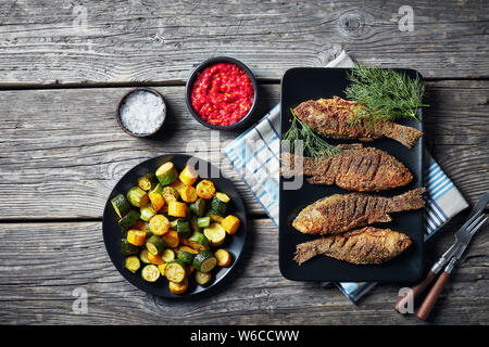 overhead view of pan-fried breaded crucian carps served on a black rectangular plate with fried zucchini slices and chili tomato sauce, flat lay Stock Photo