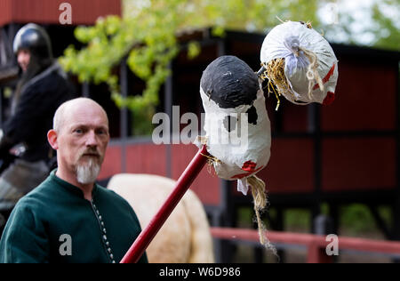 An assistant to a knight shows two heads which the knight butts with his lance during a jousting tournament at the outdoor Medieval Centre Nykøbing Falster, Denmark. Before the artificial heads was introduced in ancient times real heads from beheaded enemies or criminals was used. The Medieval Centre is a living, experimental museum which depicts the Danish Middle Ages at the 14th and 15th century. It is designed as a typical Danish market town with craftsmen, a harbour with ships and boats, and a market place. The Centre employees and volunteers are dressed in costumes of the period, they liv Stock Photo