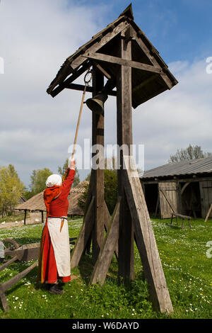 A woman rings the church bell in the little medieval village, Sundkøbing, at the outdoor Medieval Centre in Nykøbing Falster, Denmark. The Medieval Centre is built around the village Sundkøbing at a fjord as it was around the year 1400. Houses are authentically designed and built with the people who populate the village, men, women, children, peasants, craftsmen, warriors and knights etc dressing and work as they would back in 1400.  Canons and large trebuchets can be seen firing missiles, knights in armour on horseback competing with lances and swords, dyers colouring textiles with plants and Stock Photo