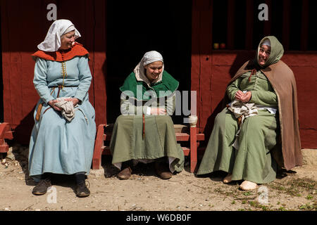 Three women in traditional costume are chatting at the little medieval village, Sundkøbing, at the outdoor Medieval Centre in Nykøbing Falster, Denmark. The Medieval Centre is built around the village Sundkøbing at a fjord as it was around the year 1400. Houses are authentically designed and built with the people who populate the village, men, women, children, peasants, craftsmen, warriors and knights etc dressing and work as they would back in 1400.  Canons and large trebuchets can be seen firing missiles, knights in armour on horseback competing with lances and swords, dyers colouring textil Stock Photo