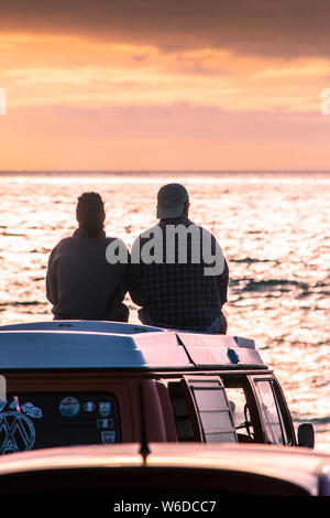 A couple sitting on top of their VW Volkswagen Campervan watching a spectacular sunset at Fistral in Newquay in Cornwall. Stock Photo