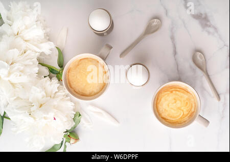 Boiled eggs in ceramic egg cups, two cups of coffee with ceramic spoons on background of beautiful white peony. Breakfast concept. Top view. Stock Photo