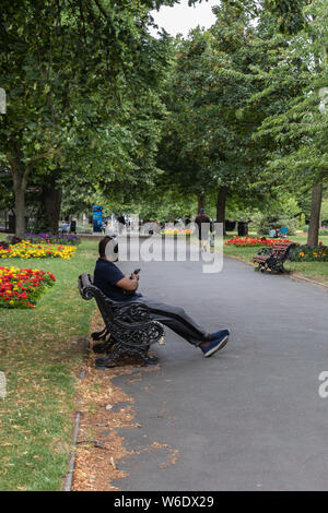 A black man sat on a bench relaxing in the park wearing headphones holding his mobile phone or sell phone Stock Photo