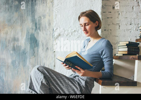 Young woman reading book on stairs in the loft art work studio room, slow life Stock Photo