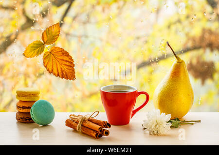 Macaroons, cup of coffee, cinnamon sticks, pear and dry leaf on window glass with water drops in the blurred background. Fallen leaf and rain drops on Stock Photo