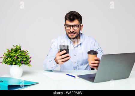 Young businessman in shirt sits in office at table and uses smartphone and drinks coffee on desk with laptop. Stock Photo