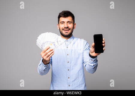 Excited man in casual t-shirt holding lots of money in dollar currencys and cell phone in hands isolated over white wall Stock Photo