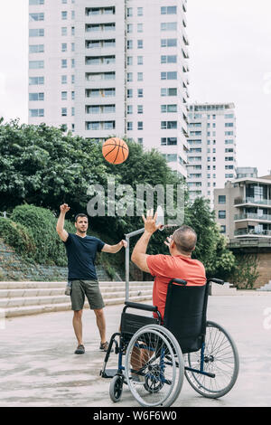 joyful handicapped man in wheelchair playing basketball with a friend with ball, concept of adaptive sports and physical activity, rehabilitation for Stock Photo