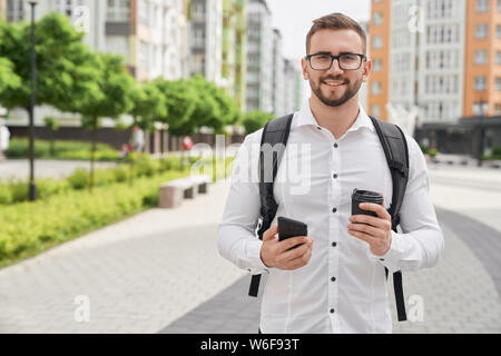 Positive, young bearded man holding phone and black coffee cup, looking at camera, smiling. Handsome freelancer posing with backpack on background of sleeping area in city. Stock Photo