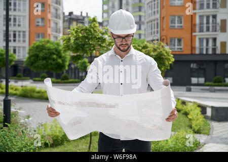 Young engineer in hardhat holding architectural construction drawings on paper on background of modern residential development. Concept of new apartments buildings. Stock Photo