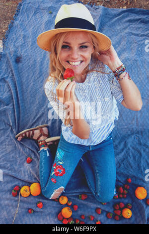 Happy young woman in hat eating strawberry on picnic in nature Stock Photo