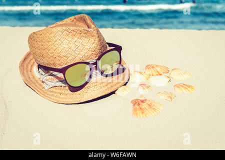 Vintage summer wicker straw beach hat and sun glasses on the sand with seashells, tropical sea background. Stock Photo
