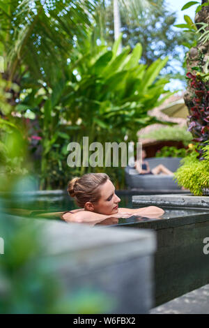 Pretty peaceful woman relaxing outdoors in sunny and lush stone swimming pool of luxurious hotel during tropical holiday in Bali Stock Photo