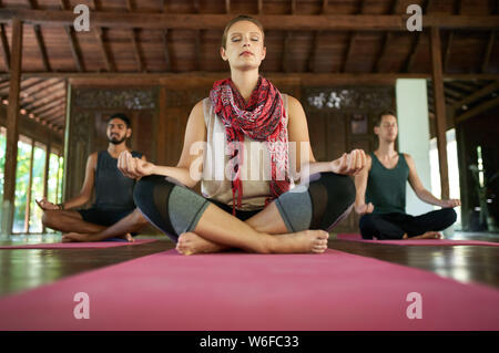 Mindful woman teaching meditation to two multi-ethnic men in lotus pose on yoga mats in traditional temple in Bali Indonesia Stock Photo