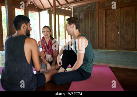 Female teacher discussing course with diverse male yoga and meditation students on yoga mats in traditional Indonesian temple Stock Photo