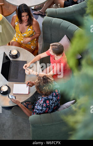 Candid shot of three diverse millennial friends talking over coffee with laptop computer at bright cafe serving fair-trade coffee Stock Photo
