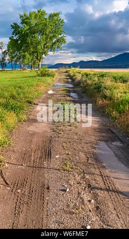 Vertical frame Dirt road on a grassy field with lake valley and mountain in the distance Stock Photo