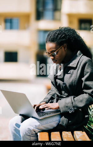 Smiling young African female university student sitting on a campus bench working on a laptop while preparing for an exam Stock Photo