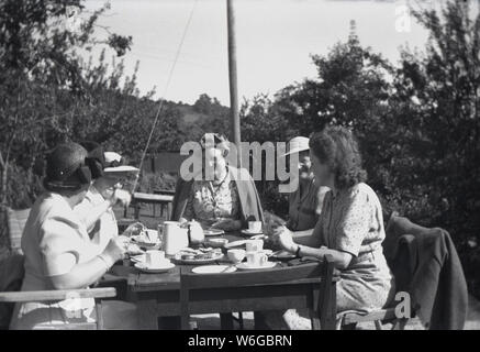 1950s, historical, summertime and a group of well-dressed English ladies wearing hats enjoying themselves, having tea and scones and good coversation sitting at a wooden table outside, England, UK. Stock Photo