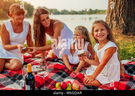 Family having picnic by summer river at sunset. Mother, grandmother and kids eating cake. Three denerations spending time together Stock Photo