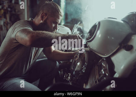 Handsome bearded man repairing his motorcycle in the garage. A man wearing jeans and a t-shirt. Stock Photo