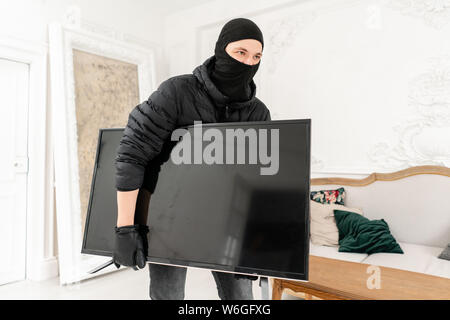 The burglar commits a crime in Luxury apartment with stucco. Thief with black balaclava stealing modern expensive television. Stock Photo