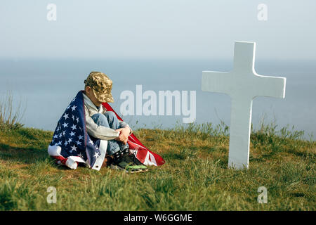 a young boy in a military cap, covered by the flag of the united states sitting on the grave of his deceased father. May 27th memorial day Stock Photo
