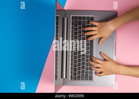 Kid hands typing on laptop computer keyboard, top view, pink and blue background Stock Photo