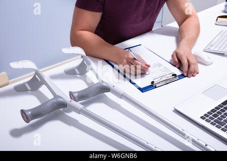 Disabled Male Patient Filling Insurance Claim Form Over Desk With Crutches Stock Photo