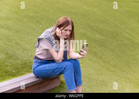 Young sad woman in a summer park sits on a bench and uses a smartphone Stock Photo