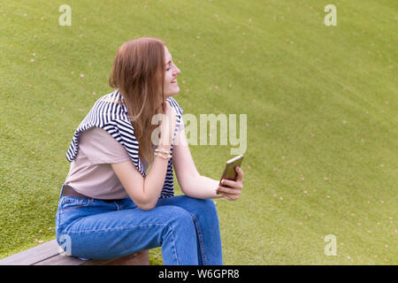 Handsome smiling woman with mobile phone sits on a park bench on a sunny day Stock Photo