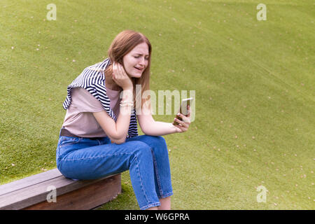 Portrait of a beautiful upset young blonde holding a mobile phone while sitting in a park on a sunny day Stock Photo
