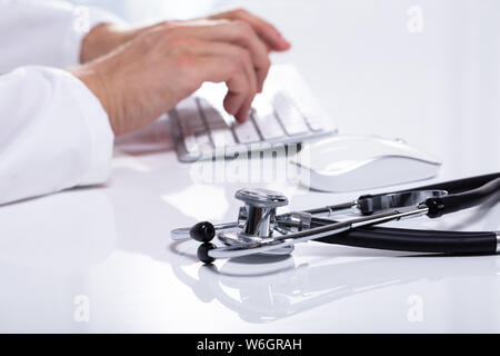 Close-up Of Stethoscope On White Desk Besides Doctor Typing On Wireless Keyboard Stock Photo