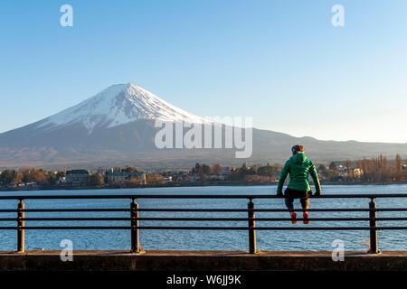 Young woman sitting on a railing, view over Lake Kawaguchi, back volcano Mt. Fuji, Yamanashi Prefecture, Japan Stock Photo