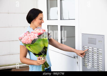 Happy Young Woman Holding Bouquet Pressing Button Of Intercom To Enter Home Stock Photo