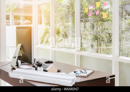 Office desk workspace stuff with computer, notepad, blueprint and coffee cup mouse notepad shot at room on tree background and sunlight. Stock Photo