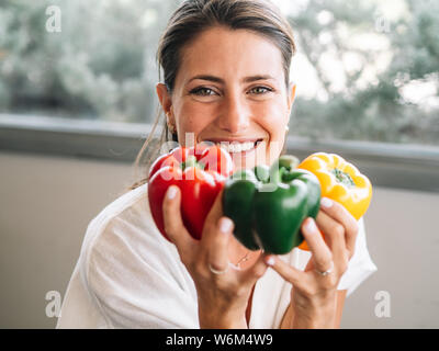 Caucasian girl sitting in her kitchen showing three big colored peppers. Green, red and yellow. Stock Photo