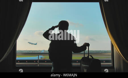 A young guy looks at the plane taking off from the window of his hotel room and leaves with a suitcase for boarding.