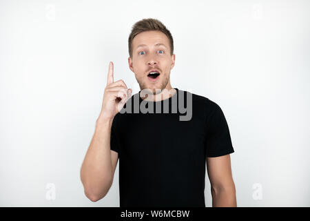 handsome young surprised man wearing black t-shirt pointing with his finger up isolated white background Stock Photo