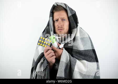 handsome young sick man wrapped checkered plaid holds pills and nasal spray looks pityingly on isolated white background Stock Photo