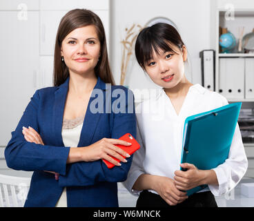 Portrait of two successful businesswomen standing with phone and clipboard in modern office Stock Photo